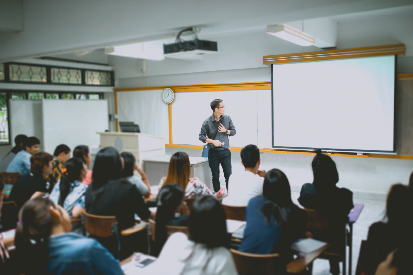 Instructor presenting to students in a classroom, standing beside a whiteboard and large projection screen while students sit at desks facing the front.