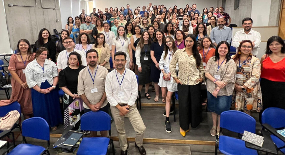 A large group of people stands together for a photo in a tiered lecture hall with grey concrete walls and fluorescent lighting. Most participants are wearing name tags around their necks. In the foreground, there are bright blue chairs. 